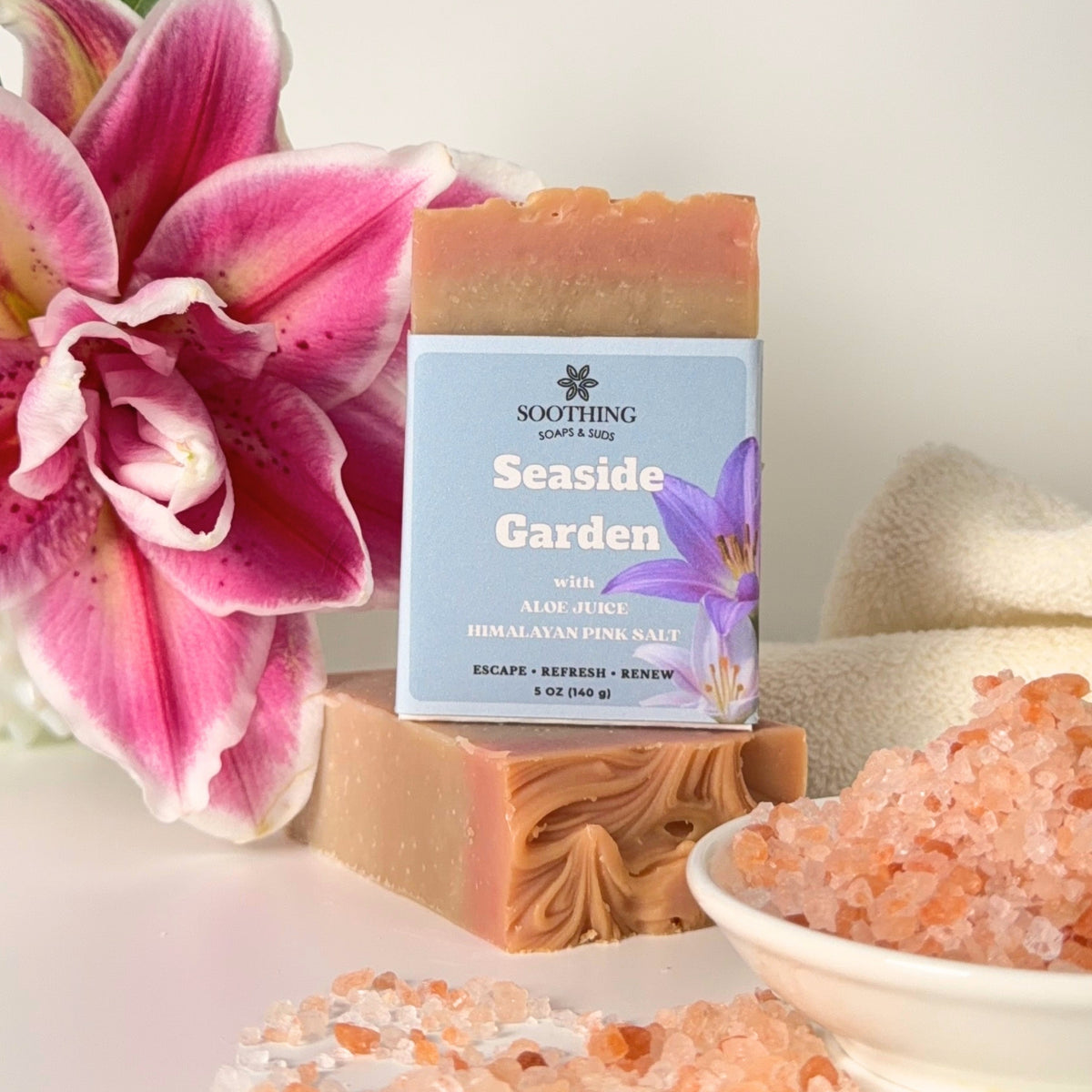 A bar of soap labeled 'Soothing Seaside Garden' stands on a white surface, styled with a pink lily flower, pink Himalayan salt, and a folded beige towel in a bright, clean setting.