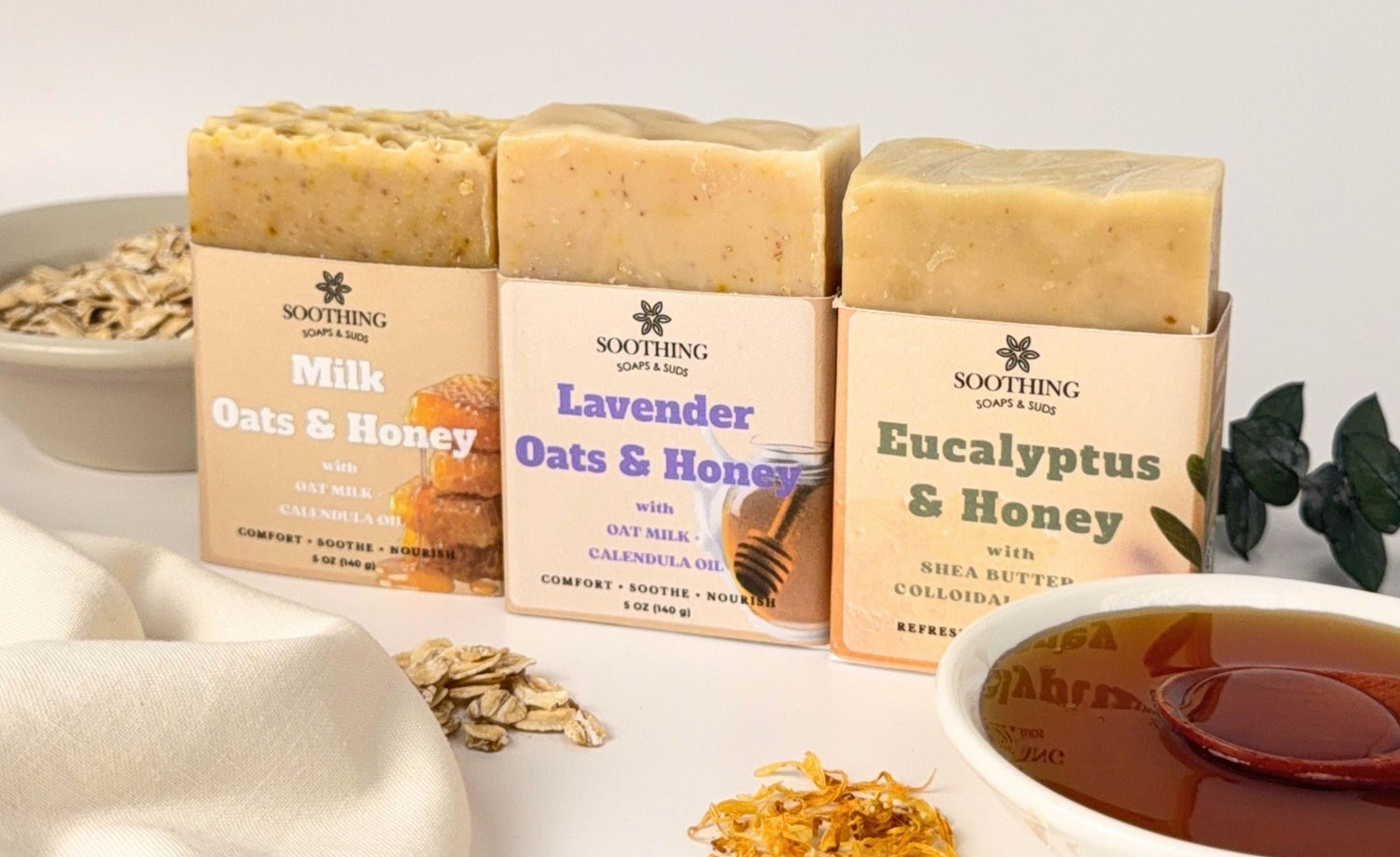 Three bars of handmade soap in their paper packaging, displayed on a white surface with natural ingredients and a bowl of honey around them. The soaps are labeled 'Soothing Body Soaps' with flavors 'Milk Oats & Honey', 'Lavender Oats & Honey', and 'Eucalyptus & Honey'.