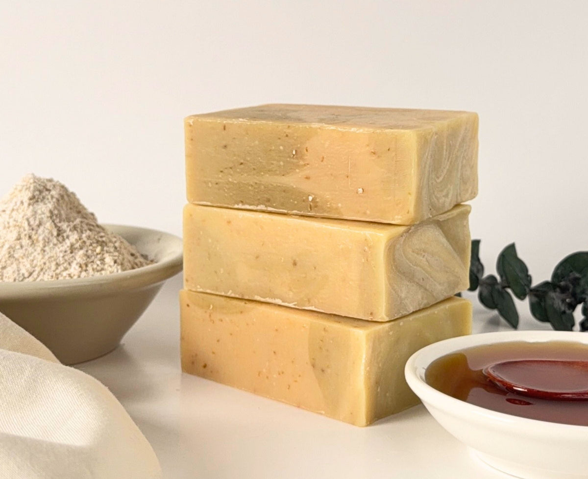 Three stacked rectangular bars of handmade soap in a warm light beige color with visible speckles, displayed on a white surface with bowls of ingredients and a cloth nearby.